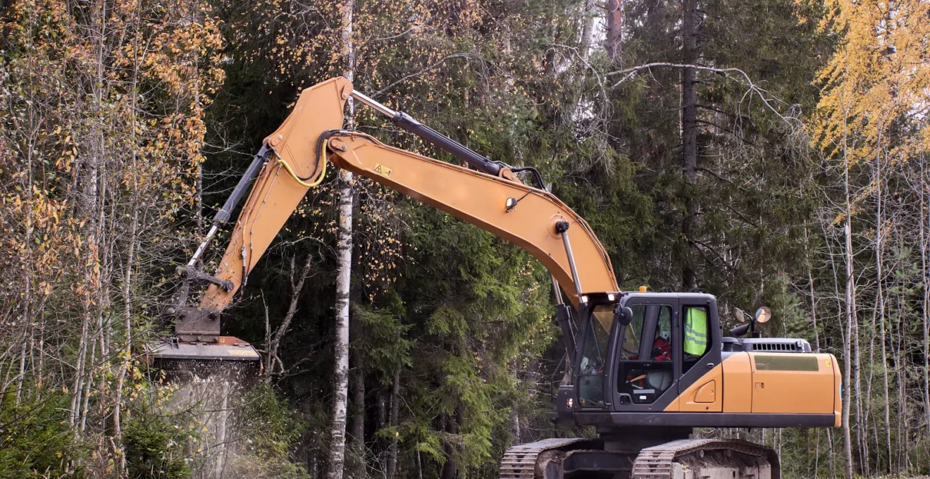 Un opérateur manœuvre une excavatrice chenillée équipée d'une tête de broyage sur un terrain en pente couvert de broussailles.